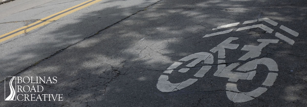 A logo for a bike path painted on the asphalt street. A double yellow line appears in the corner of the photo.